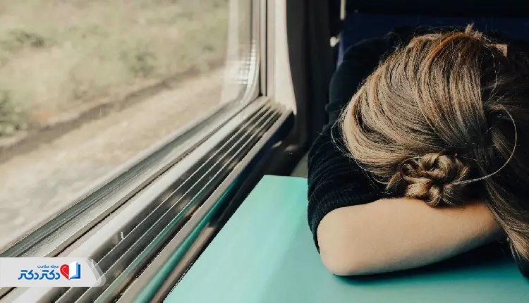 A tired woman resting her head on a table near a window, possibly experiencing fatigue related to anemia, which may be detected through an MCH test